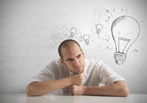 A man sits at a table, resting his chin on his fist and looking thoughtful—perhaps pondering questions to ask someone about their career. Above his head are sketches of light bulbs, with one large bulb symbolising inspiration.