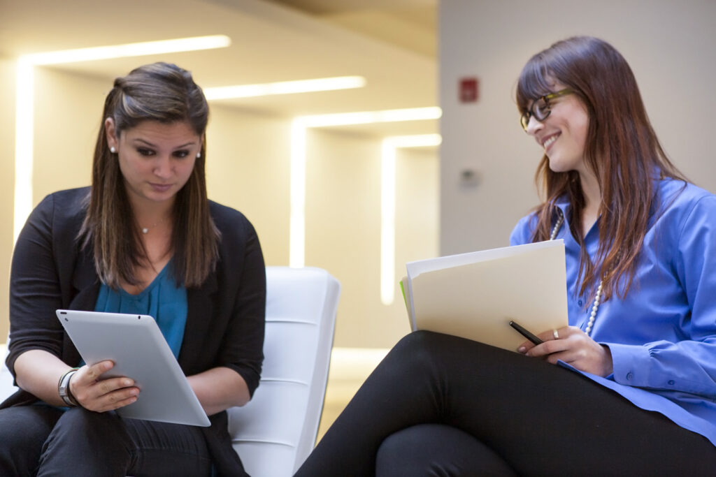 Two women sit together indoors; one looks at a tablet while the other, holding a folder and pen, smiles at her. They appear to be engaged in a friendly, informal career development discussion.