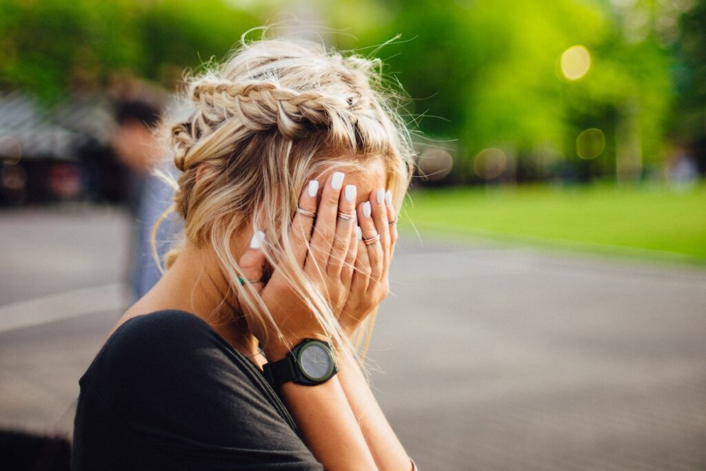 A person with blonde hair styled in plaits covers their face with their hands outdoors, perhaps feeling impostor syndrome. They wear a black shirt, a wristwatch, and have white polished nails, with greenery and blurred people in the background.