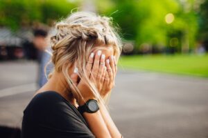 A person with blonde hair styled in plaits covers their face with their hands outdoors, perhaps feeling impostor syndrome. They wear a black shirt, a wristwatch, and have white polished nails, with greenery and blurred people in the background.