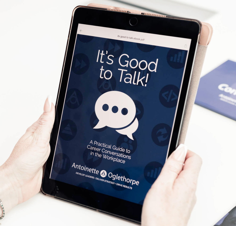 A person holds a tablet displaying an e-book cover titled "It's Good to Talk! A Practical Guide to Career Conversations in the Workplace" by Antoinette Oglethorpe. The cover is blue with a speech bubble icon.