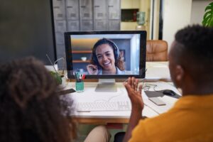Two people sit at a desk and video chat with a smiling woman on a computer screen, discussing Managing Careers in a Hybrid Workforce. Papers, keyboard, and office supplies are visible on the desk in this modern office setting.