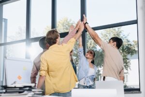 A group of five young adults stand in an office by large windows, smiling and high-fiving to celebrate teamwork and measuring the success of career development. Sunlight streams in, with a flipchart full of sticky notes visible nearby.