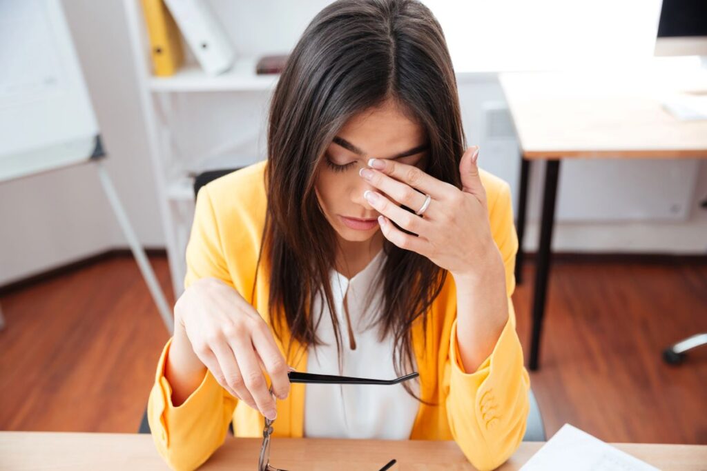 A woman wearing a yellow jacket sits at a desk, holding her glasses in one hand and rubbing her eyes with the other, appearing tired—perhaps reflecting on talent development mistakes in an office setting.