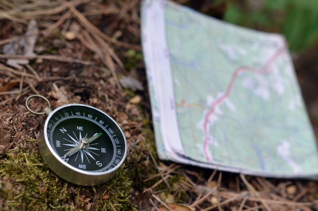 A close-up of a compass resting on the ground beside a folded, slightly blurred map in an outdoor setting, symbolising navigation or orienteering in nature and the journey towards career clarity.