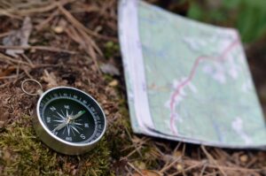 A close-up of a compass resting on the ground beside a folded, slightly blurred map in an outdoor setting, symbolising navigation or orienteering in nature and the journey towards career clarity.