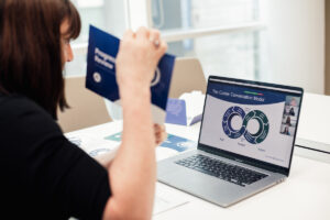 A person sits at a desk holding a pamphlet, looking at a laptop screen displaying a video call with slides titled