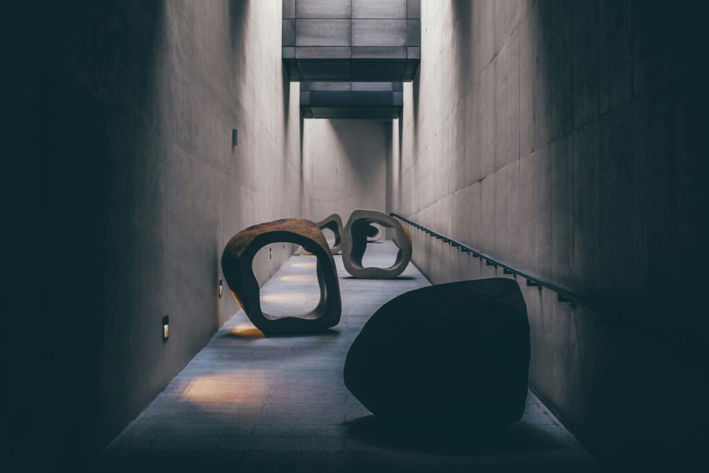 A dimly lit corridor with smooth concrete walls features abstract, hollow stone sculptures placed along a gently sloping ramp, symbolising overcoming obstacles as they cast soft shadows on the floor.