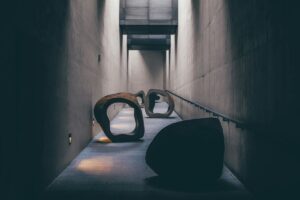 A dimly lit corridor with smooth concrete walls features abstract, hollow stone sculptures placed along a gently sloping ramp, symbolising overcoming obstacles as they cast soft shadows on the floor.