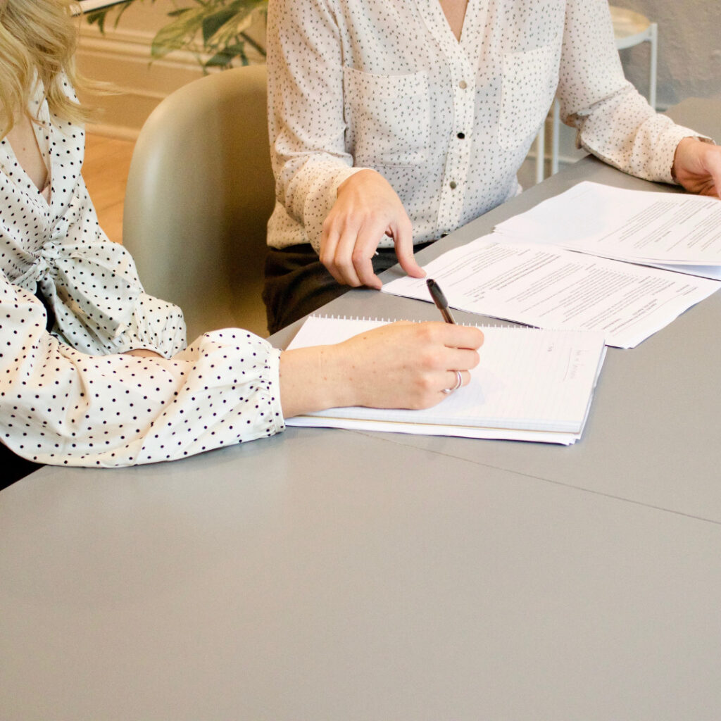 Two people in white, polka-dotted tops sit at a table with documents and notebooks, discussing and writing notes together in a bright office setting as they explore the essential skills and qualities of managers.