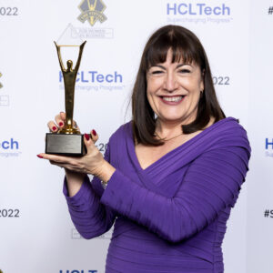 A smiling woman in a purple dress holds a gold trophy, celebrating her Stevie Awards win in front of a step-and-repeat backdrop featuring HCLTech and Stevie Awards logos.