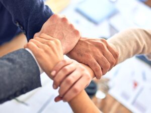 Four people clasp each other's wrists, forming a connected circle with their hands, symbolising teamwork, trustworthiness, and unity. The background is blurred with office documents and supplies visible.
