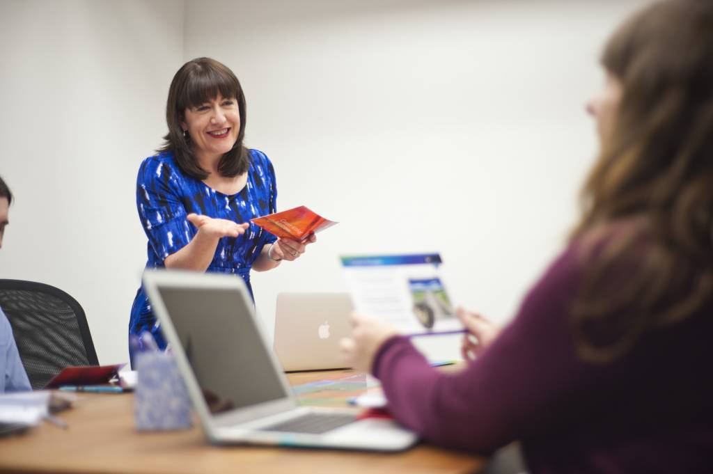 Antoinette Oglethorpe in a blue dress smiles and gestures while leading career conversations round a table with laptops and papers, engaging with a person in the foreground.