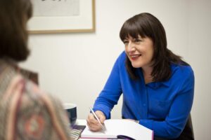 A woman in a blue shirt smiles whilst writing in a notebook, facing another person in the foreground. They appear to be engaged in a friendly conversation, reflecting the collaborative spirit of good executive coaching in an office setting.