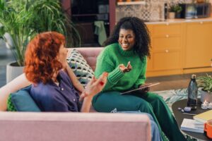 Two women sit on a pink sofa in a cosy, colourful living room, smiling and talking. One holds a tablet and gestures while the other claps, both appearing cheerful as they discuss ways to improve emotional intelligence together.