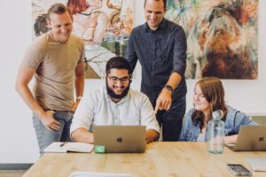 Four smiling people gather around a table with laptops and notebooks, demonstrating emotional intelligence as they collaborate in a bright office adorned with colourful abstract art. One person points at a laptop screen while others look on, engaged and supportive.