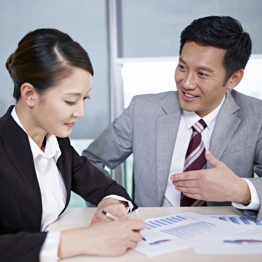 A woman in business attire writes on a document whilst a man in a suit sits beside her, smiling and gesturing, as they discuss performance appraisals with charts and graphs on the table in front of them.