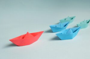 Several paper boats on a light blue surface, with one red boat leading ahead of four blue boats that follow behind in a staggered formation.
