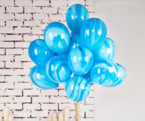 A bunch of shiny blue balloons are gathered together in front of a white brick wall, with several white candles placed on the floor in the background.