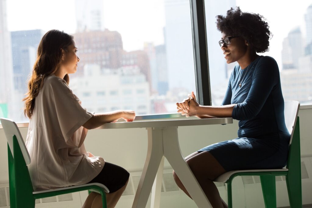 Two women sit opposite each other at a table by a large window, engaged in conversation about being a great mentee, with a vibrant cityscape visible in the background.