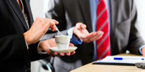 Two people in business attire are having a discussion at a desk. One stirs coffee while the other gestures over documents, demonstrating how to have a career conversation in a professional setting.