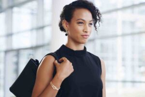 A woman with short curly hair, wearing a sleeveless black top and pearl earrings, stands indoors with a black bag over her shoulder, boosting confidence as she looks ahead. The background is bright and blurred.
