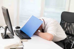 A man sitting at an office desk, appearing stressed or exhausted, rests his head on his arms beneath a blue folder. Surrounded by a computer, phone, office supplies, and a Gartner report, he seems overwhelmed by work.