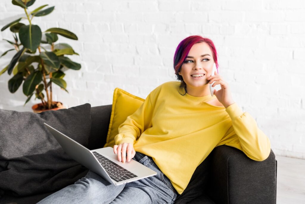 A Gen Z individual with pink hair, wearing a yellow jumper and jeans, sits on a dark sofa with a laptop on their lap, smiling whilst talking on a mobile phone. A yellow cushion and a plant are visible in the background.