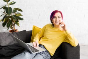 A Gen Z individual with pink hair, wearing a yellow jumper and jeans, sits on a dark sofa with a laptop on their lap, smiling whilst talking on a mobile phone. A yellow cushion and a plant are visible in the background.