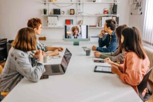 Four people sit at a conference table with laptops and notepads, participating in a video call with a person displayed on a monitor—reflecting the collaborative nature of today’s hybrid workplace in a bright, modern office.
