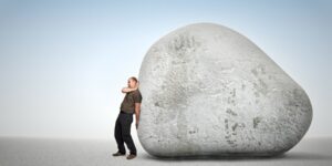 A man in dark clothing stands with his back against a huge grey boulder, looking tired and burdened as he supports the weight of the rock on his shoulders—showing why it’s important to help your employees take ownership.