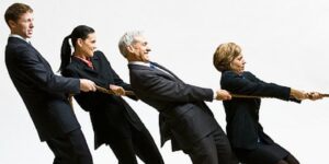 Four people in business suits, two men and two women, stand in a row pulling on a rope as if in a tug-of-war, symbolising resistance to change as they lean back and work together against a plain white background.