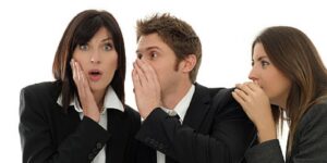 Three people in business attire whispering to each other, with surprised and curious expressions, as they discuss creating a coaching culture, all set against a plain white background.