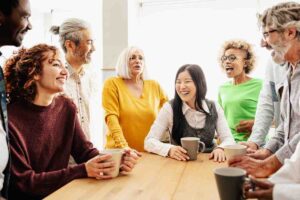 A diverse, multi-generational workforce gathers around a table, smiling and laughing together whilst holding coffee mugs in a bright room.