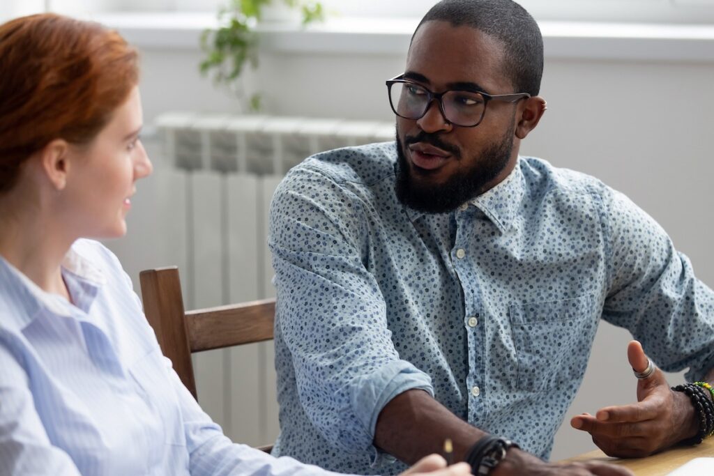 A man wearing glasses and a patterned shirt gestures while speaking to a woman with red hair during a conversation about their organisations mentoring programme in a bright, casual indoor setting.