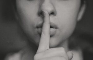 A close-up, black-and-white photo of a person holding a finger to their lips in a gesture signalling silence or secrecy, subtly capturing the weight of self-doubt. The image is slightly blurred and the background is out of focus.