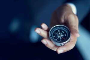 A person in a suit holds a silver compass in their hand, the needle pointing southeast—symbolising strategies for success. The blurred, dark background draws attention to the hand and compass as the image’s focal points.