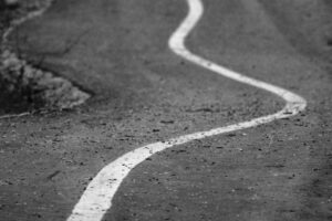 A close-up of a winding, curvy white line on a paved road or path, surrounded by scattered debris and blurred edges, evokes the abstract, zigzag pattern of a non-linear career path.
