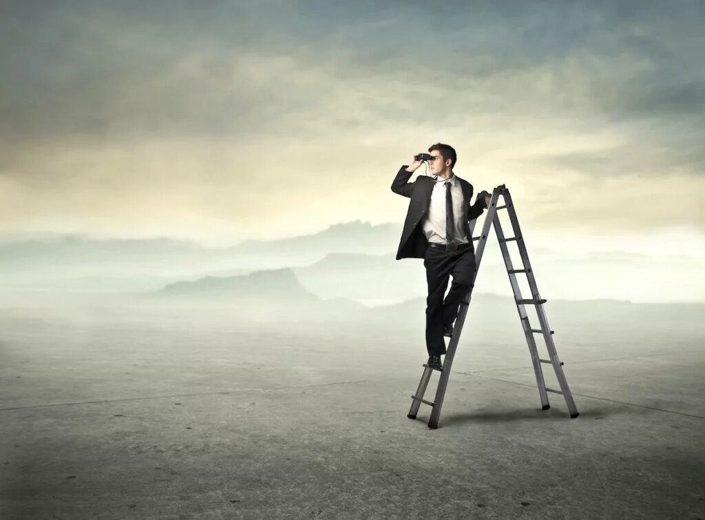 A man in a suit stands on a ladder in an empty, expansive landscape, looking into the distance through binoculars under a cloudy sky, symbolising a search for the future of work.