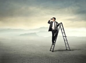 A man in a suit stands on a ladder in an empty, expansive landscape, looking into the distance through binoculars under a cloudy sky, symbolising a search for the future of work.