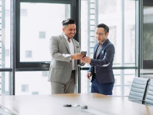 Two men in business suits stand by a large window in a modern office, smiling and looking at a mobile phone together—perhaps sharing tips on how to better understand your manager. An empty meeting table and chairs are in the foreground.