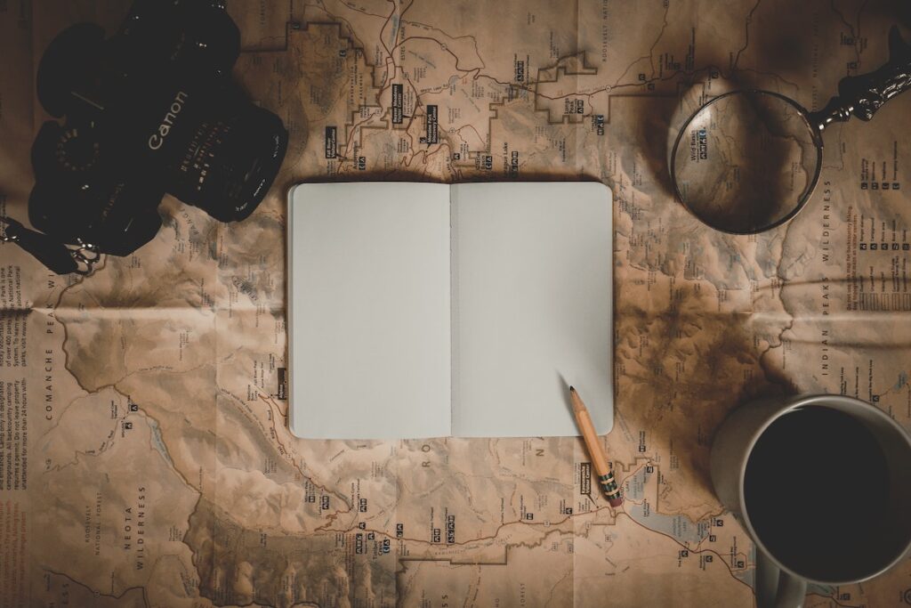 An open blank notebook lies on an old map, surrounded by a camera, magnifying glass, pencil, and coffee—creating a vintage travel planning scene that suggests being well-prepared for a career conversation or new adventure.