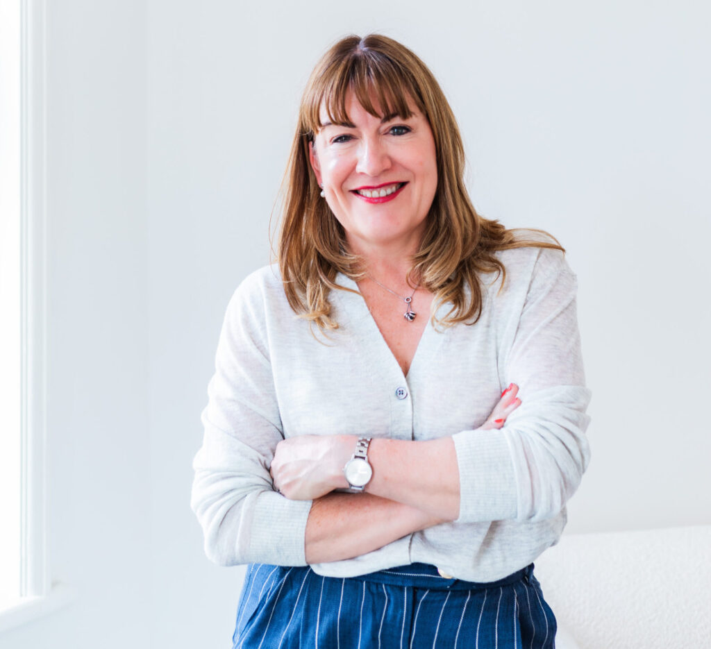 Antoinette Oglethorpe profile picture, she has straight brown hair and a fringe smiles while standing with arms crossed. She is wearing a light cardigan, a necklace, a wristwatch, and striped trousers, against a bright, minimal indoor background.
