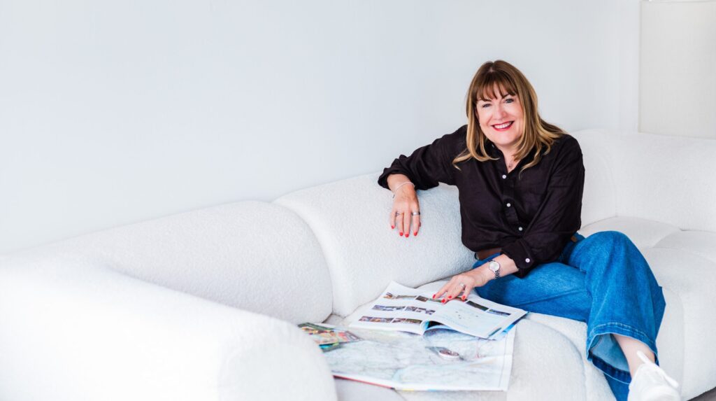 Antoinette Oglethorpe with straight, shoulder-length hair, wearing a black blouse and blue jeans, sits smiling on a white sofa. She has an open magazine and papers spread out on the cushion beside her.