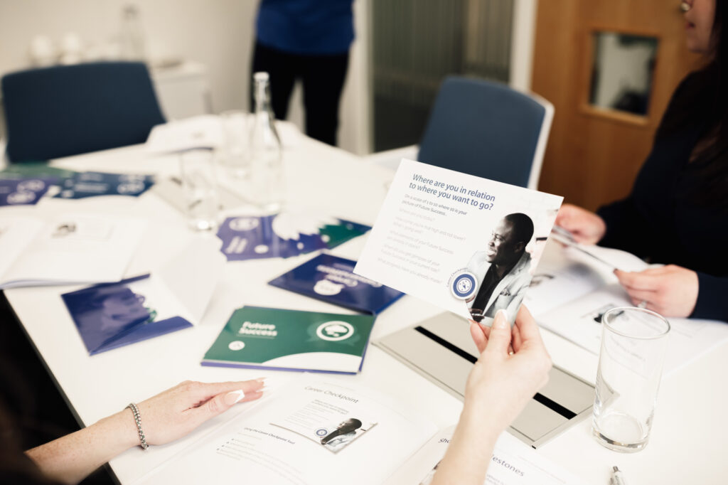 A person holds a leaflet at a table covered with brochures, booklets, and a glass of water, while another person sits nearby. The setting appears to be a meeting or workshop.