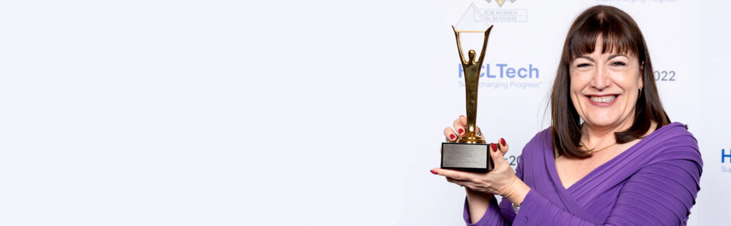 Antoinette Oglethorpe in a purple dress holds up a gold trophy against a white backdrop with event logos, celebrating her achievement.