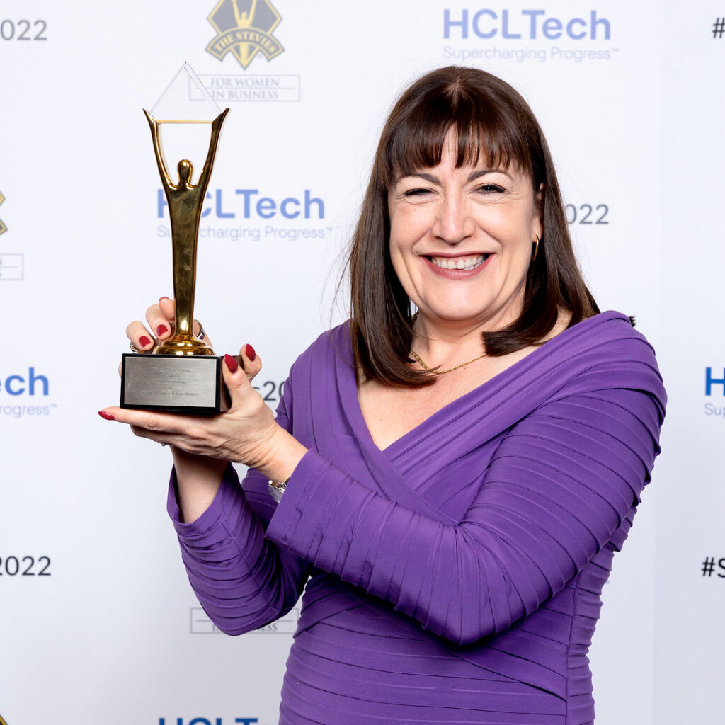 Antoinette Oglethorpe in a purple dress smiles whilst holding up a gold winners trophy. The backdrop features logos for HCLTech and the Stevie Awards for Women in Business.