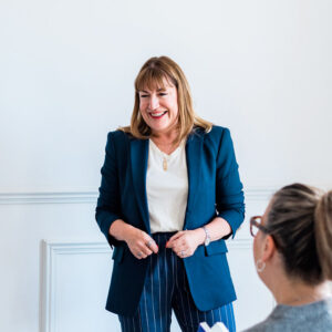 Antoinette Oglethorpe keynote speaker and global authority on career development, leadership and the future of work in a blue blazer stands and smiles whilst speaking to seated people in a bright room with a white wall.