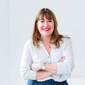 Antoinette Oglethorpe with light brown hair and fringe, wearing a light jumper and striped trousers, stands smiling with arms crossed in a bright room next to a window.
