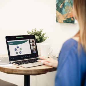 A woman sits at a small round table, attending a webinar on-demand on her laptop. A booklet, white mug, and small plant are on the table, and a painting hangs on the white wall in the background.